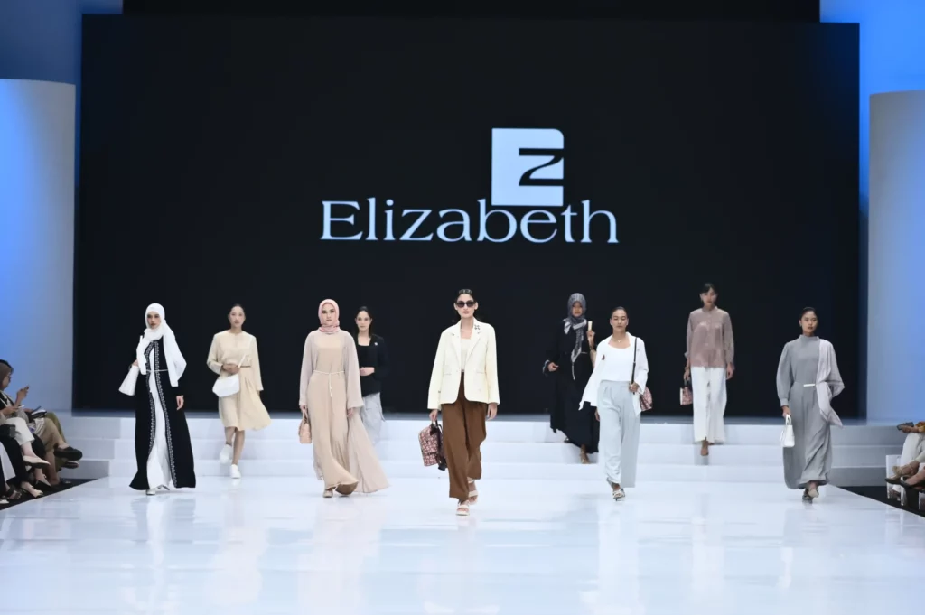 sekelompok model yang sedang berjalan di catwalk dengan latar belakang logo 'Elizabeth' pada event fashion show. Model-model tersebut memperagakan berbagai outfit yang mencakup pilihan gaya berhijab dan non-hijab dengan warna netral seperti putih, krem, dan coklat. Mereka membawa tas-tas yang nampaknya merupakan bagian dari koleksi "A Classic Ikat" oleh Elizabeth, menampilkan beragam desain seperti handbag, sling bag, dan tas bahu yang elegan. Audience tampak menyaksikan dengan antusias, menggambarkan suasana Indonesia Fashion Week 2024.