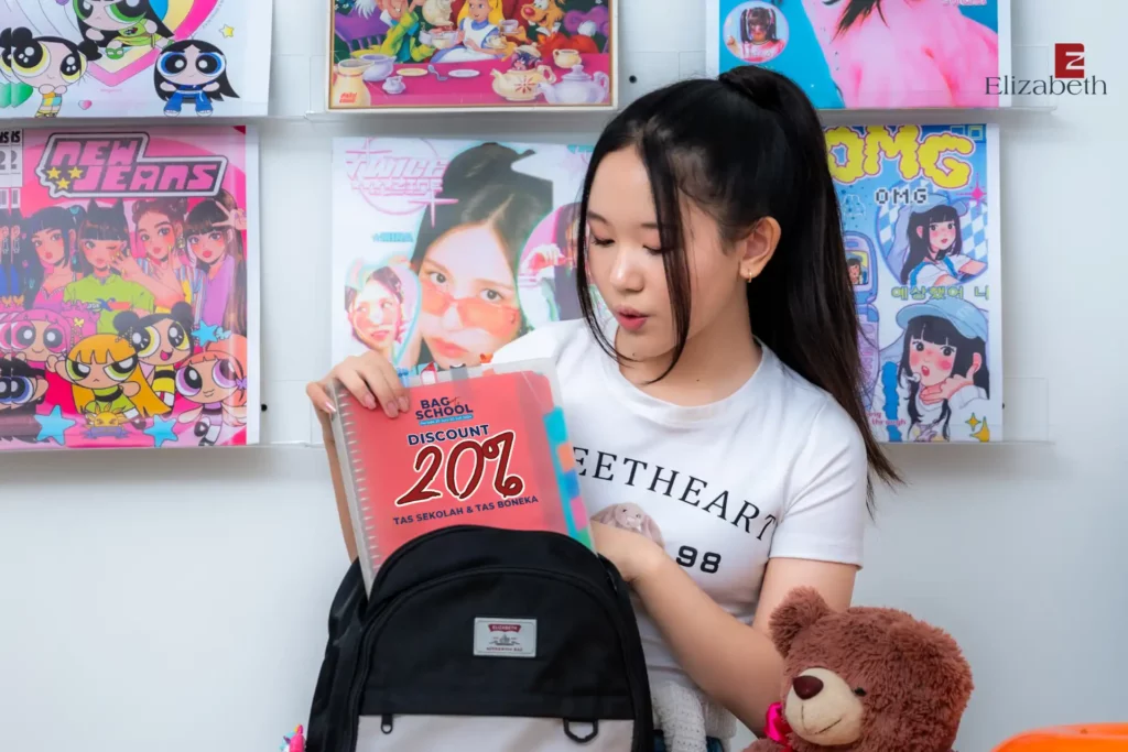 seorang anak perempuan memegang tas sekolah elizabeth sambil mengeluarkan buku dengan sampil tulisan promo bag to school elizabeth